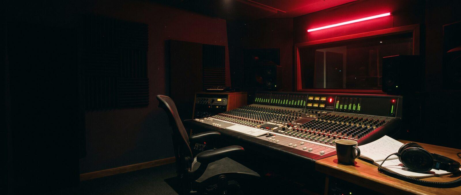 Empty Liverpool recording studio at three in the morning, analogue mixing console with green and red LED meters glowing, red neon strip on the back wall, headphones and coffee mug on the desk